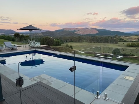 An outdoor space features a saltwater pool surrounded by smooth decking and glass fencing. A shaded area with a patio umbrella is visible, alongside reclining deck chairs. The backdrop includes rolling hills and mountains under a colorful sunset sky.