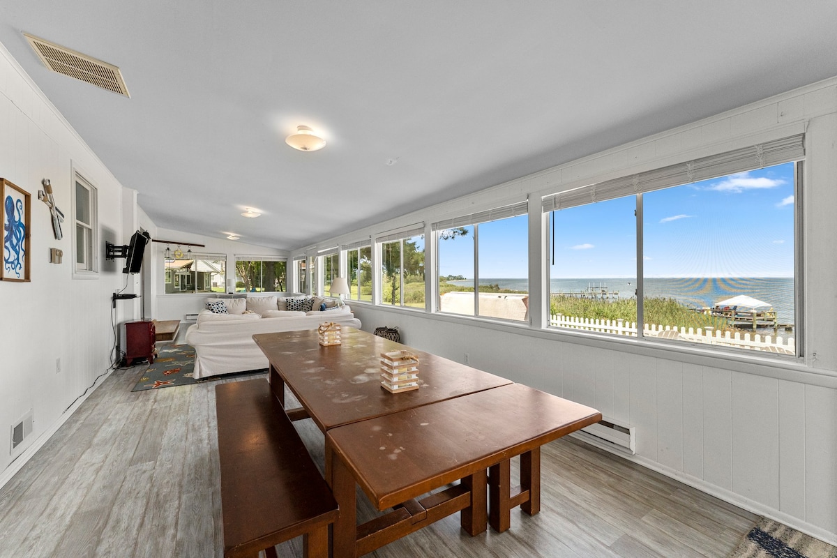 A spacious living area features large windows that provide an uninterrupted view of the beach. A wooden dining table is complemented by a casual seating space with light-colored sofas, surrounded by natural light, creating a welcoming environment.