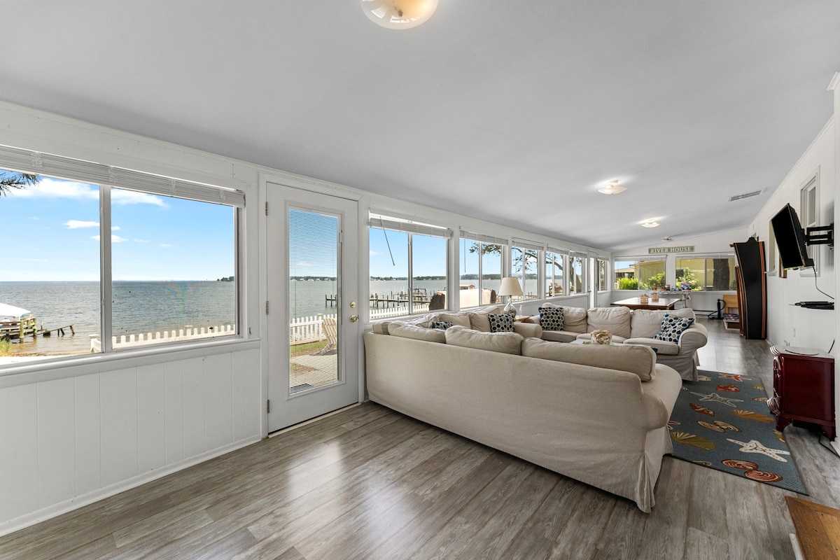 A bright living space features large windows providing views of the water and landscape. The room is furnished with a light-colored sofa and carpet that includes colorful designs. Natural light fills the area, enhancing the open and airy ambiance.