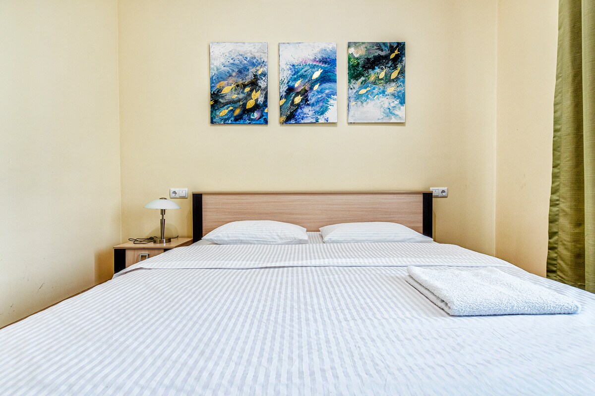 A spacious bed is centered in the room, topped with a neatly arranged blanket and a folded towel. Above the bed, three art pieces depicting aquatic scenes are mounted on the light-colored wall. Soft lighting is provided by a bedside lamp on a small wooden table.