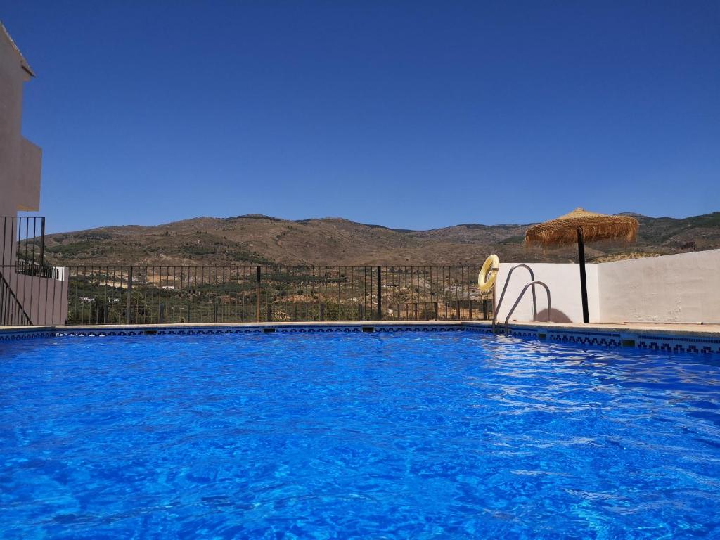 A clear blue swimming pool is shown, reflecting the bright sky above. Surrounding hills are visible in the distance, providing a scenic backdrop. A straw umbrella stands nearby, offering shade, while the area is enclosed by a low fence, enhancing the tranquil setting.