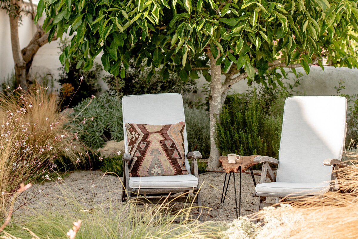 Two comfortable chairs are positioned under a leafy tree, surrounded by a garden featuring native plants. A small table holds a beverage, enhancing the serene setting. Texture is added by the cushion on one chair and the sandy ground, creating a relaxing outdoor space.