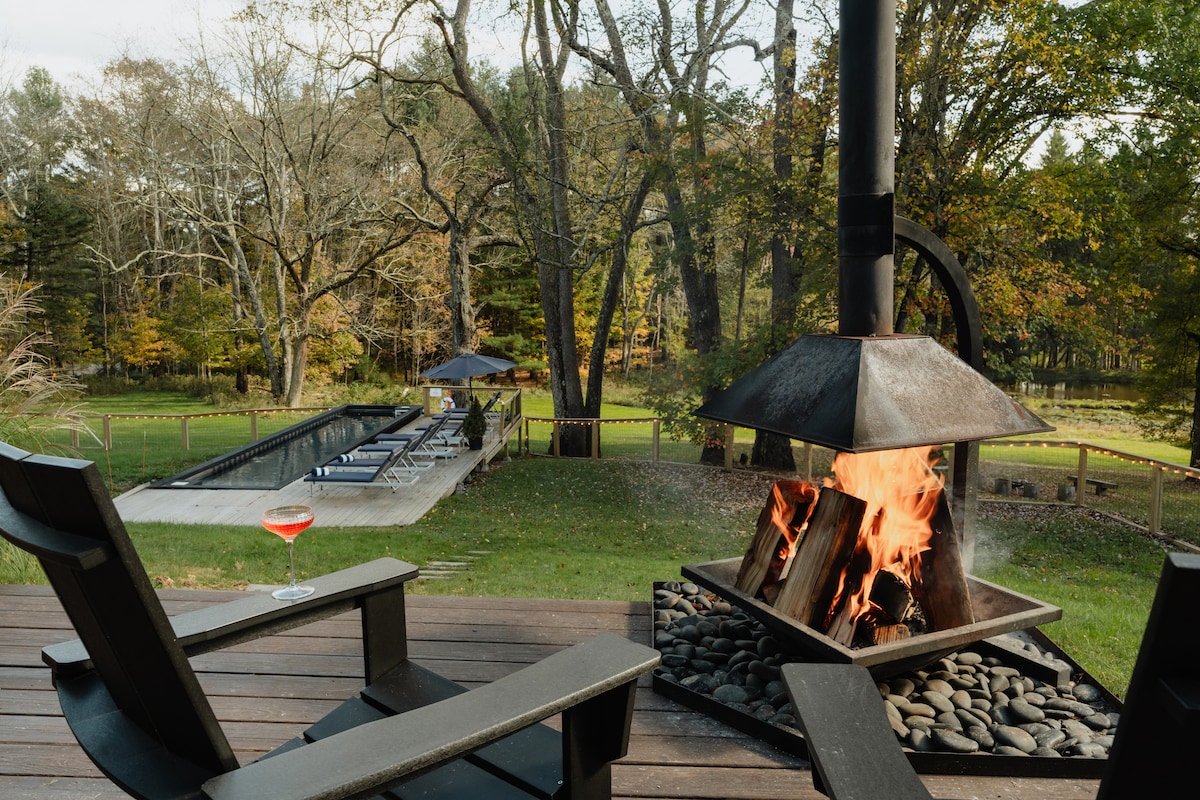 An outdoor seating area features two wooden chairs positioned near a metal fire pit, where flames flicker above a bed of stones. In the background, a heated pool surrounded by lounge chairs is visible, set against a backdrop of trees and a serene landscape.