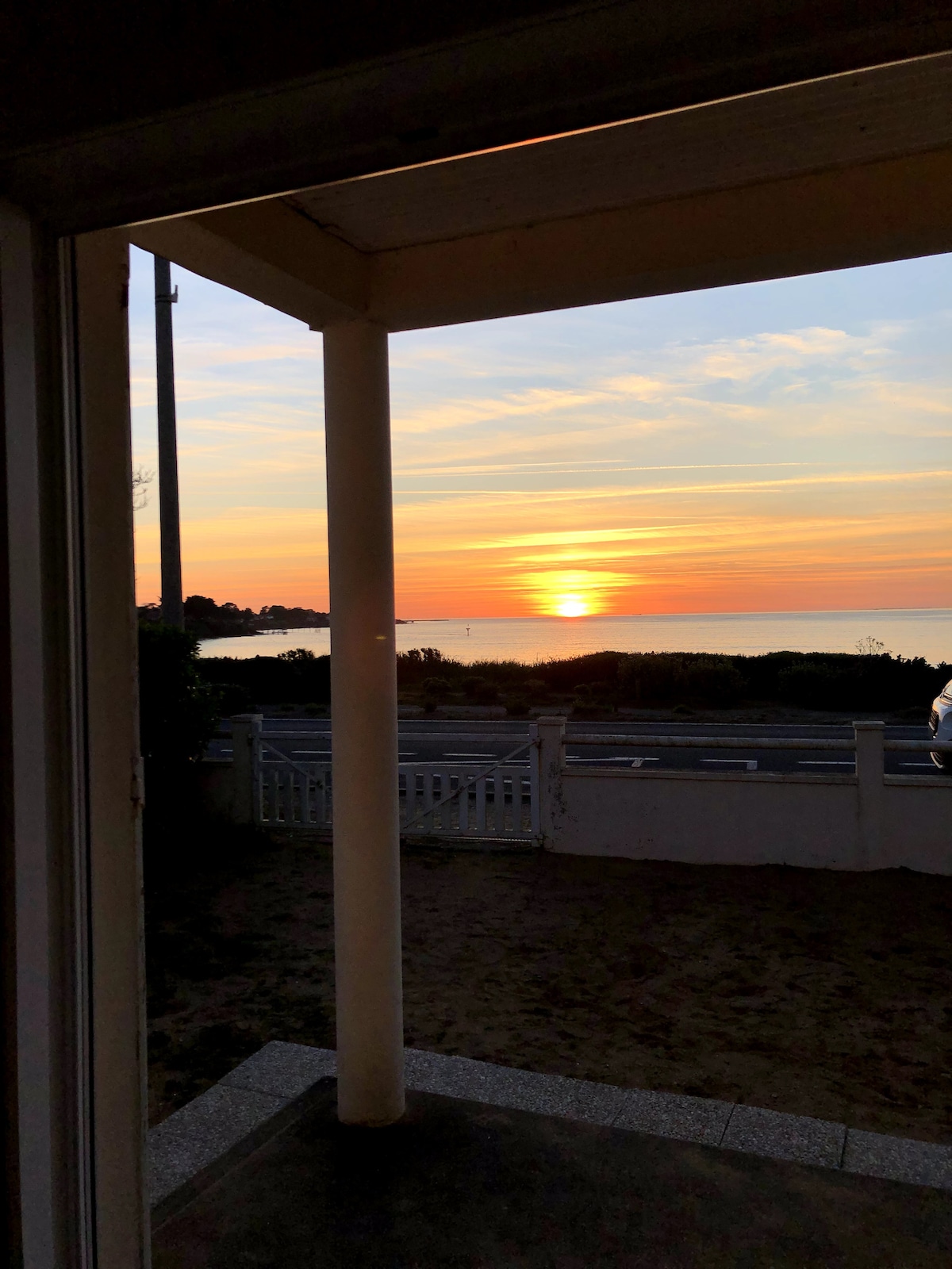 A sunset view is captured from a doorway, showcasing vibrant colors reflecting on the ocean. The horizon blends warm hues of orange and yellow with soft blue tones. A sandy area is visible in the foreground, alongside white fencing that frames the scene.