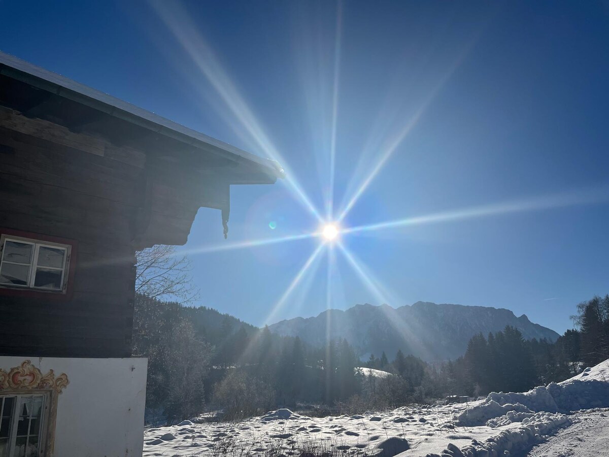 A sunlit winter landscape is showcased, with the sun casting rays over snow-covered terrain. A wooden structure with a detailed gable is visible in the foreground, alongside distant mountains framed by trees in the background.