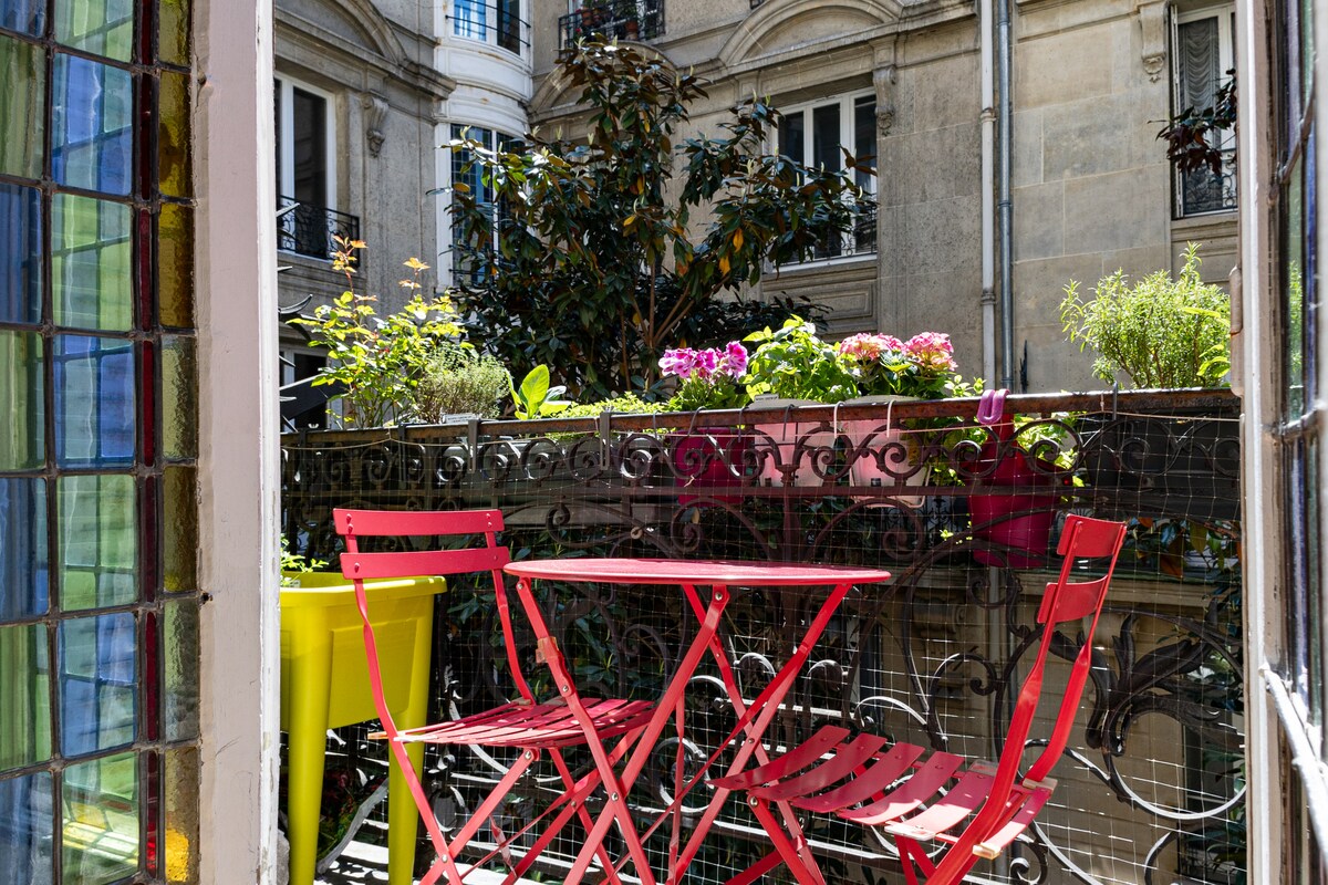 A small balcony is framed by a stained glass window, featuring a red bistro table set with matching chairs. Potted plants and colorful flowers enhance the greenery, contributing to a serene outdoor space overlooking the courtyard.