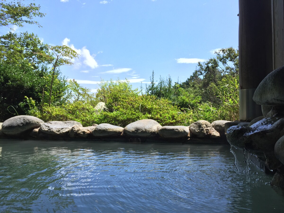 A natural hot spring bath is visible, bordered by smooth stones. The water reflects the clear blue sky, dotted with soft white clouds. Lush greenery surrounds the area, providing a serene backdrop for relaxation.