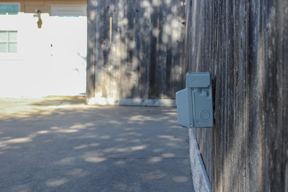 An exterior view of a wooden fence with a mounted gray electrical outlet. The scene is illuminated by natural light, creating soft shadows on the ground. A doorway can be seen in the background, providing access to the property.