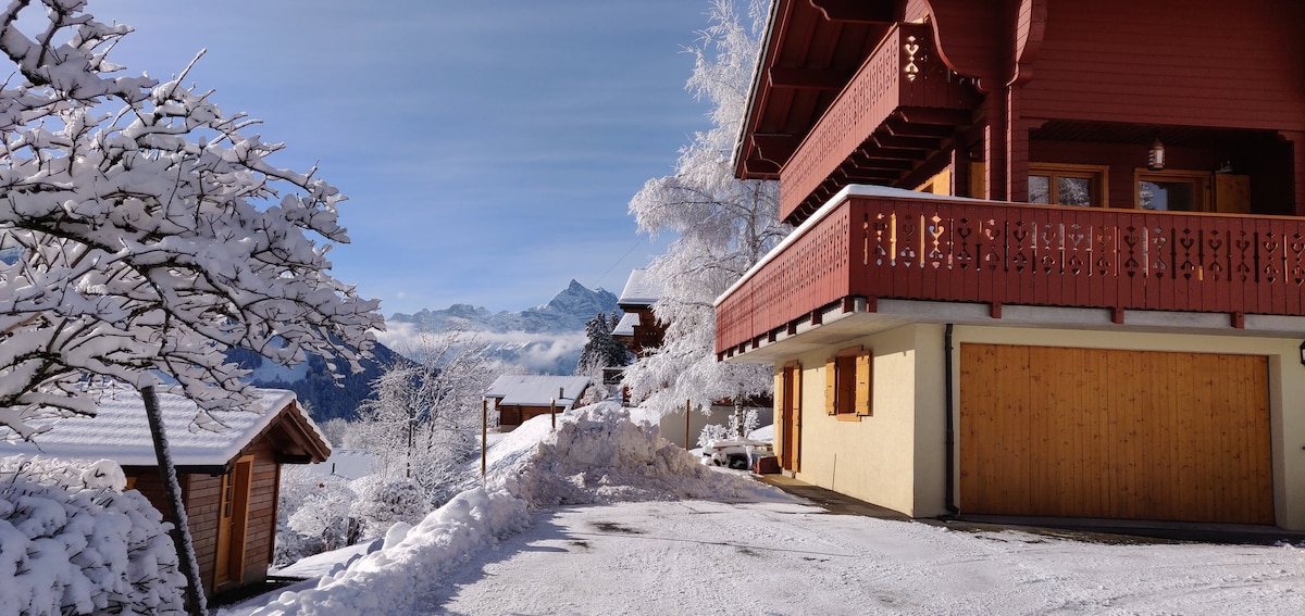 The family chalet is surrounded by a snowy landscape, showcasing striking mountain views. The wooden exterior is complemented by a balcony adorned with decorative railings. A driveway leads to a medium garage, while snow-covered trees frame the scene, highlighting the tranquil winter atmosphere.
