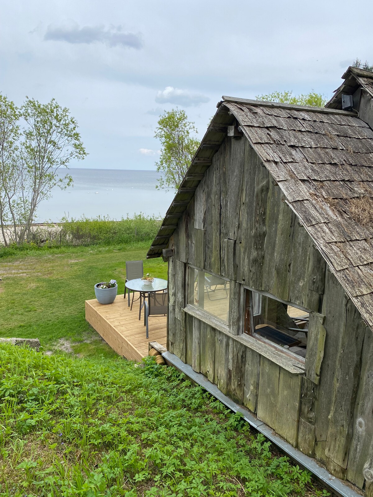 A rustic fisherman shed is positioned near the shoreline, featuring a weathered wooden exterior with a sloped roof. A wooden deck with outdoor seating overlooks the grassy area and distant sea, framed by a few trees and a cloudy sky.
