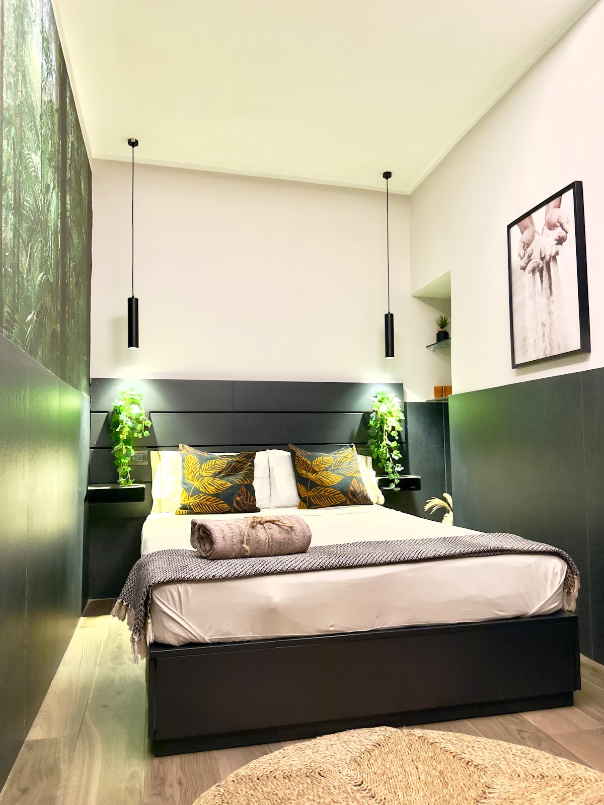 A modern bedroom features a spacious bed with decorative pillows showcasing a botanical design. Green plants are positioned on either side, complemented by stylish pendant lights. Light wood flooring adds warmth to the overall design, with a piece of wall art displayed above the bed.