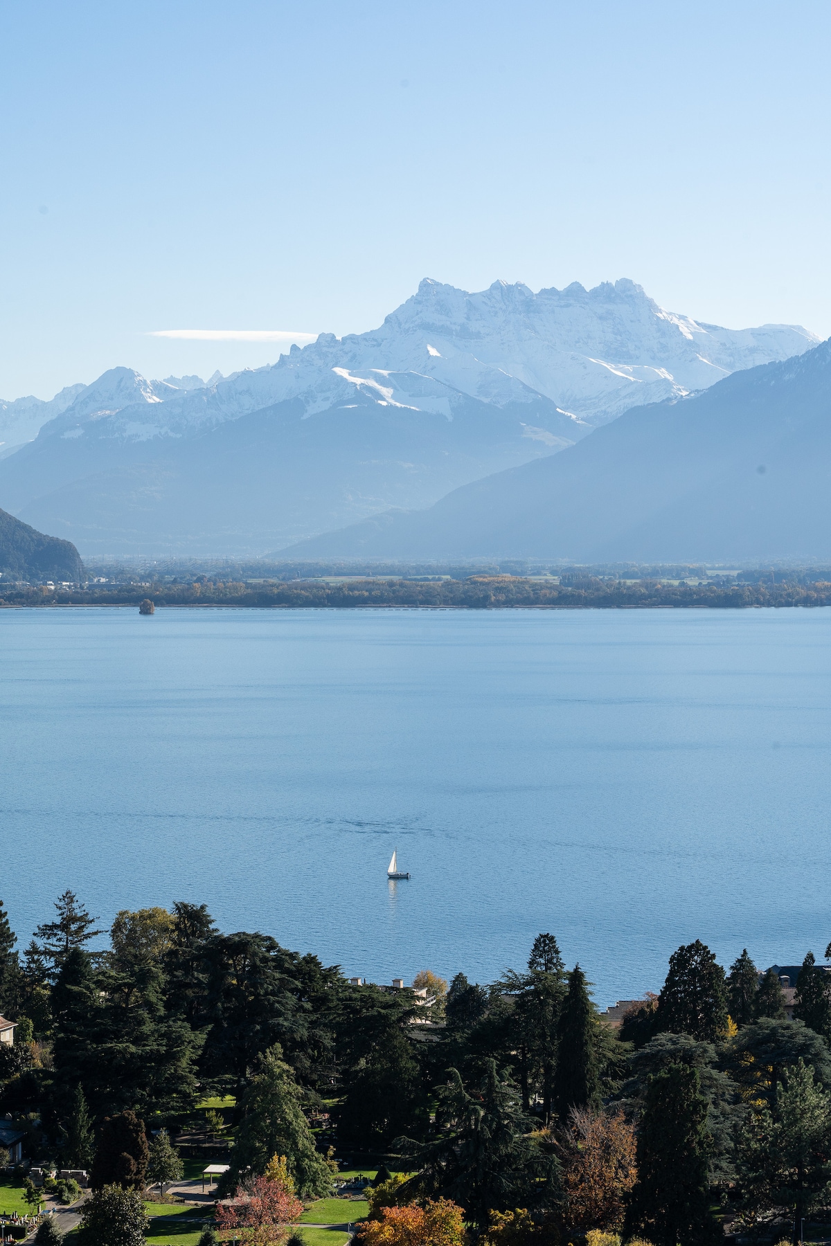 A serene view of Lake Geneva is displayed, framed by majestic snow-capped mountains in the distance. A single sailboat glides across the calm water, while lush greenery is visible along the shoreline, contributing to the scenic landscape.