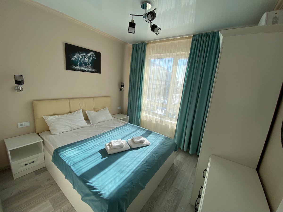 A softly lit bedroom features a spacious bed with white linens topped by a light blue blanket. Natural light filters through sheer curtains, illuminating the room's neutral tones. Two bedside lamps provide additional lighting, and a piece of wall art is displayed above the headboard.