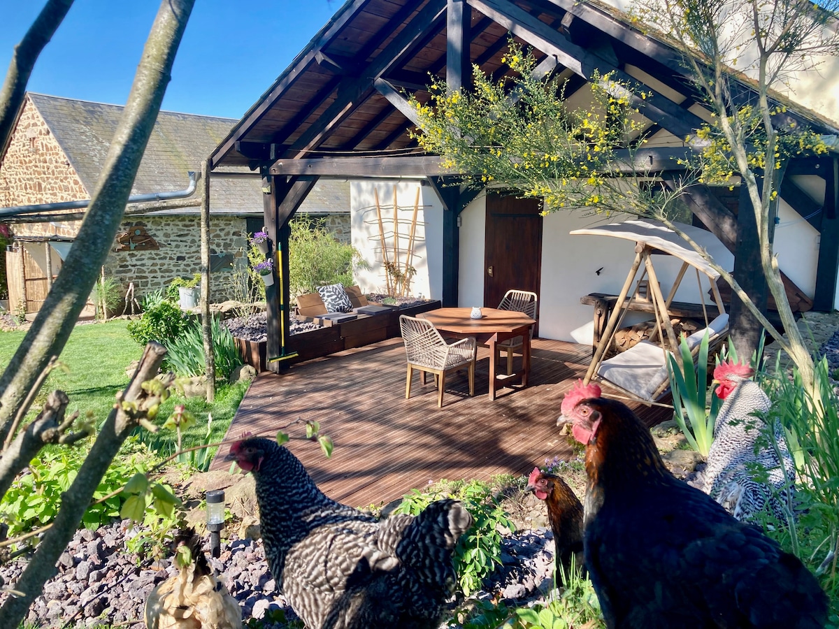 A rustic outdoor seating area is situated beneath a wooden overhang, featuring a round table surrounded by four chairs. In the foreground, chickens roam freely among greenery and flowers, enhancing the atmosphere of the peaceful garden.