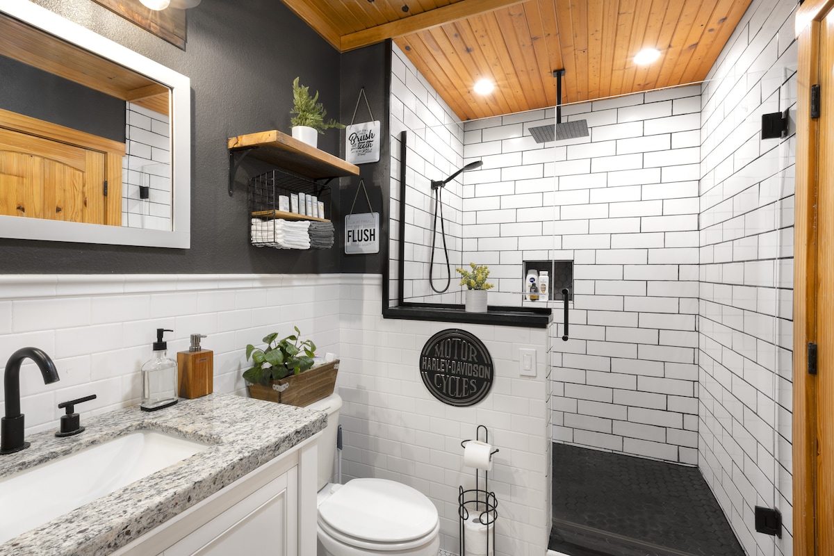 A modern bathroom features a fully tiled shower with a large rain shower head. A sleek granite countertop with a sink and a towel shelf complements the space. Decorative plants add a natural touch against the white tiled walls.