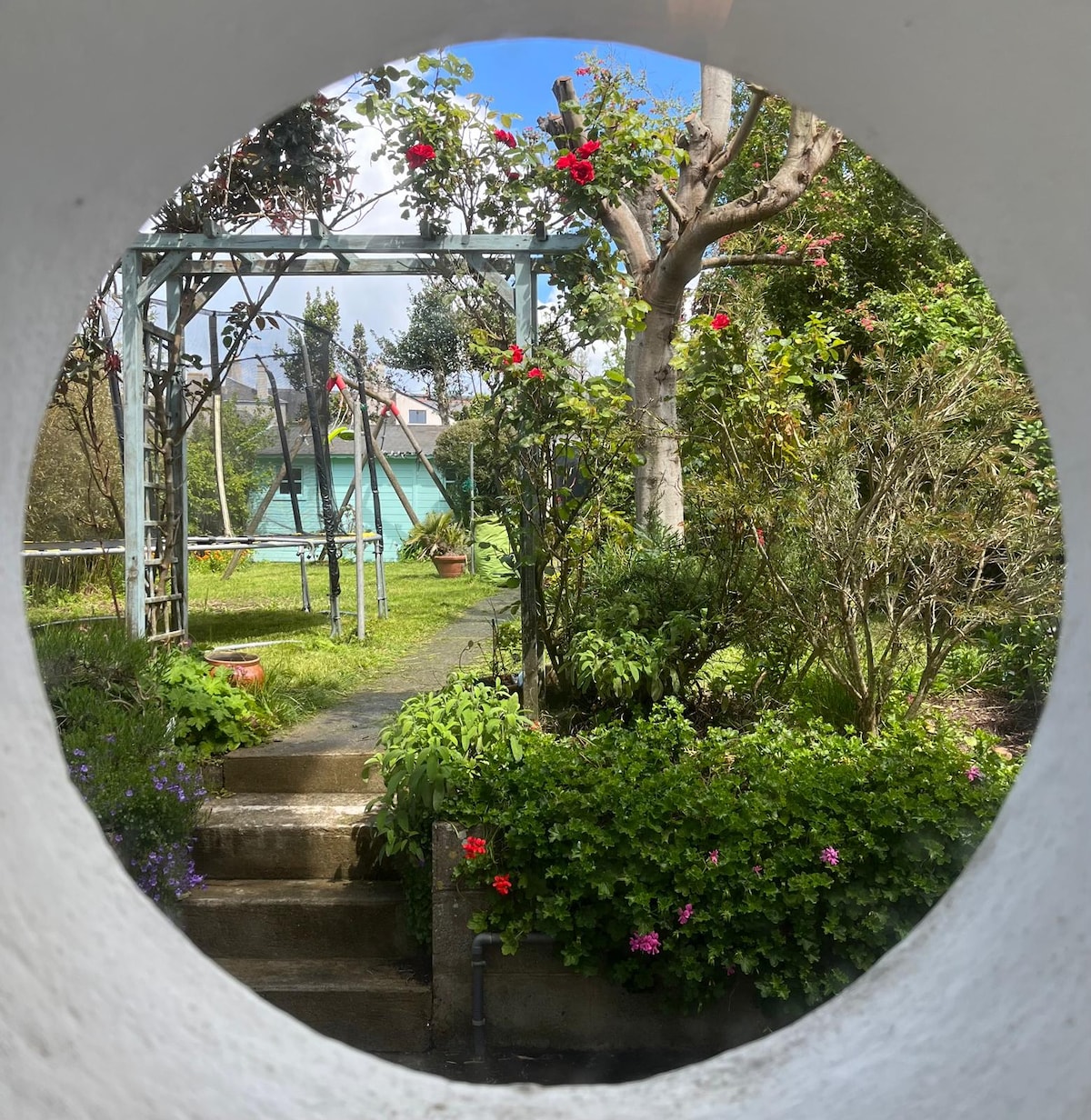 A serene garden is framed by a circular opening, showcasing a lush landscape filled with vibrant flowers and greenery. Steps lead into the garden, where a swing set is visible amongst the trees and plants, inviting relaxation and outdoor enjoyment.