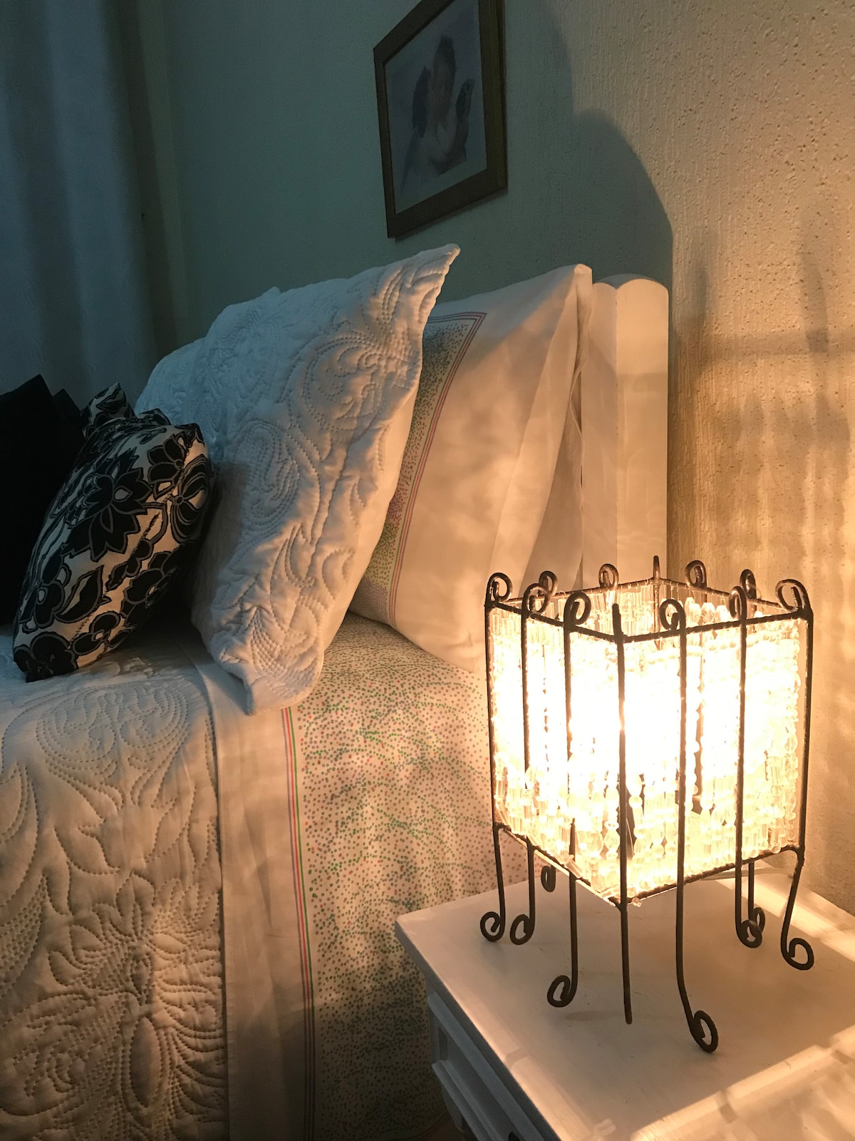 A spacious bed is dressed in crisp white linens and a textured quilt. A decorative black and white pillow adds contrast. Soft light emanates from a stylish lamp with a metal frame and glass accents, creating a warm ambiance in the room.