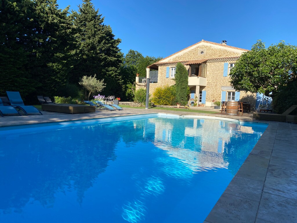 A clear blue swimming pool reflects the surrounding greenery and the 18th-century building. Sun loungers are arranged along the poolside, providing a space for relaxation. Lush trees and hedges frame the garden area, enhancing the tranquil environment.