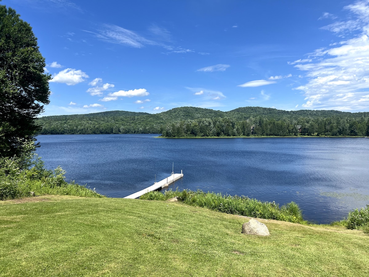A serene view of Lake Harris features a gently sloping green lawn leading to the water's edge. A white dock extends into the lake, framed by lush greenery and distant hills under a clear blue sky.