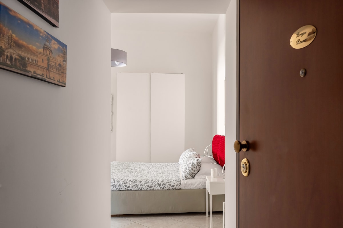 A glimpse into the apartment is provided through the open door, featuring a single bed adorned with floral bedding and a simple bedside table. A red accent blanket is placed on the bed, and a wardrobe is visible in the background.