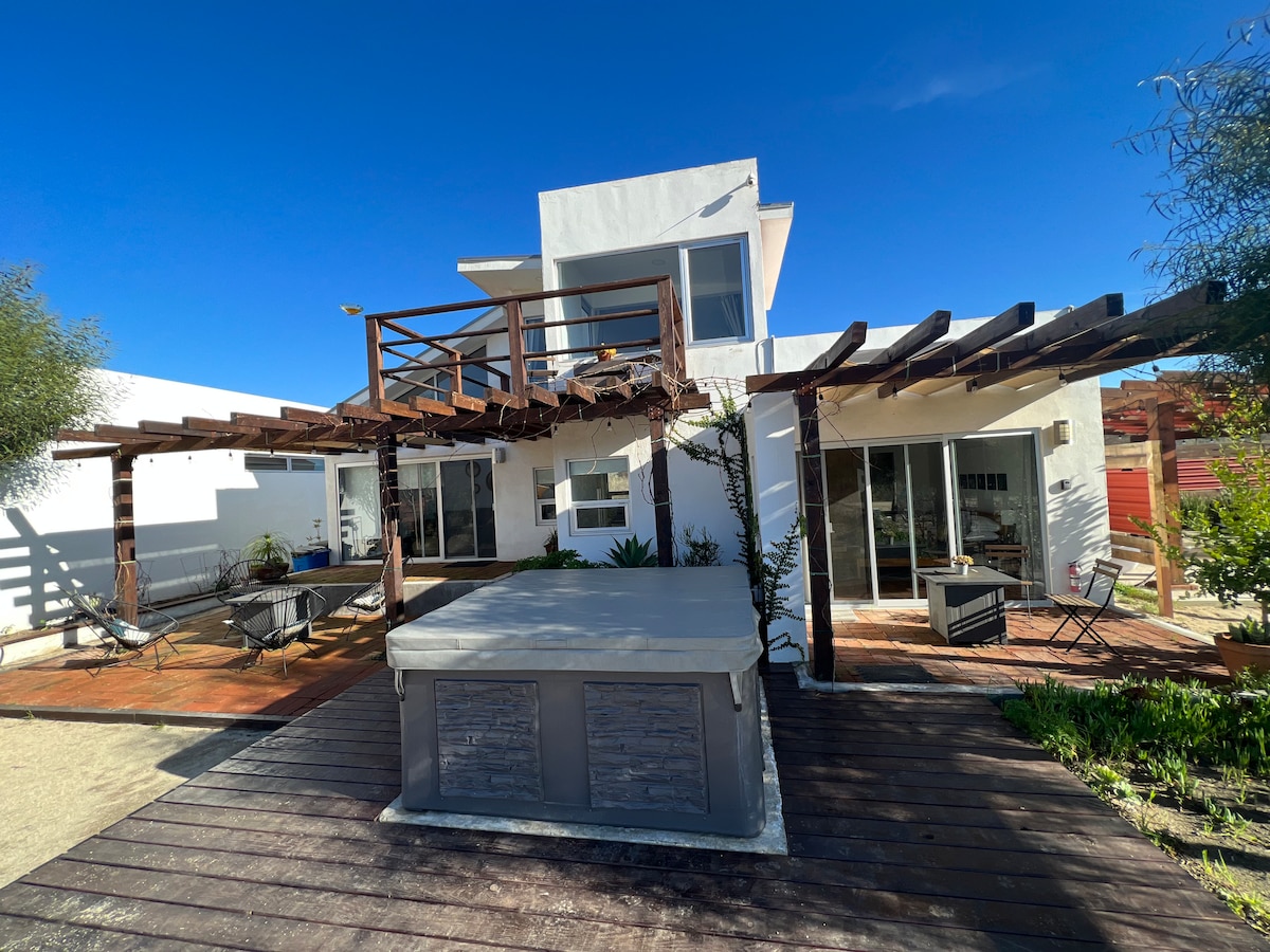 A modern, two-story house features a spacious outdoor area with a jacuzzi positioned on a wooden deck. Large glass doors provide access to the inviting interior. The surrounding landscape includes greenery and minimalist seating arrangements, creating a peaceful environment.