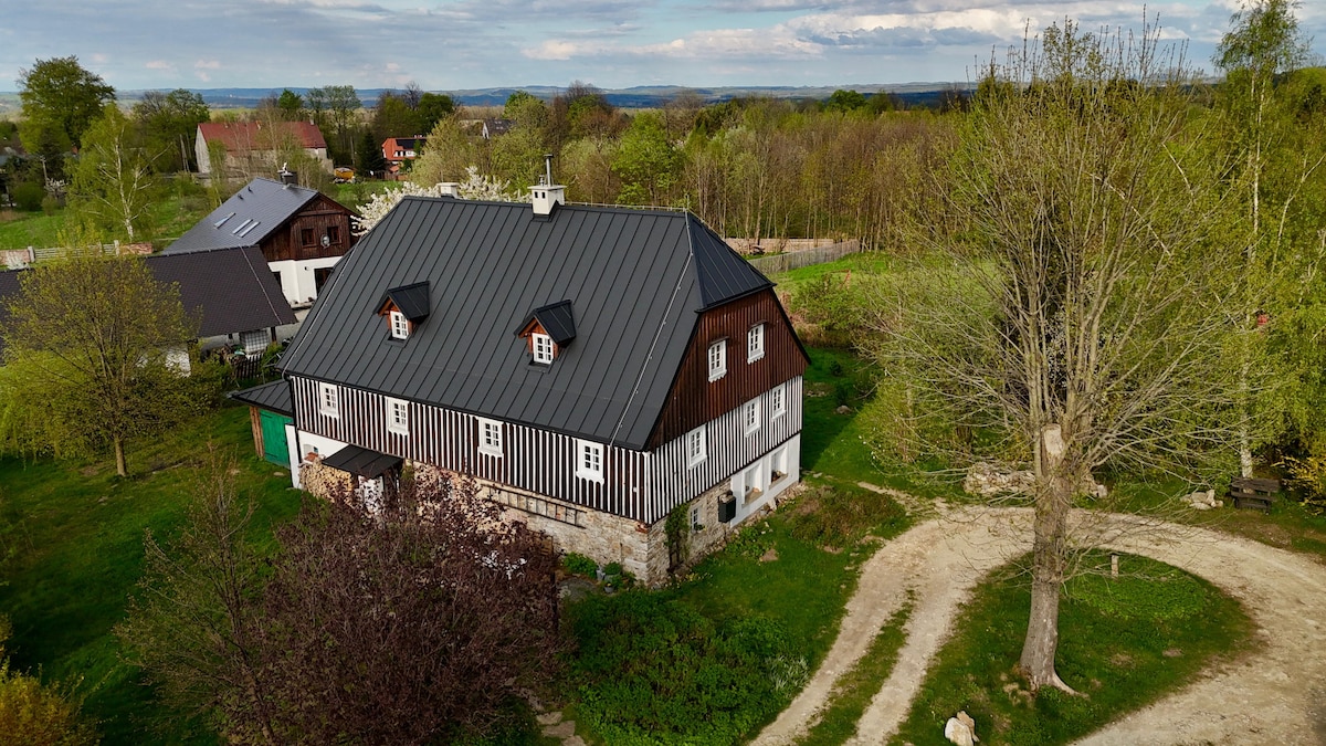 The exterior of a historic two-hundred-year-old house is presented, featuring a black and white facade with a gabled roof. Surrounding greenery and a winding dirt path are visible, with distant hills providing a scenic backdrop. The area shows a rustic charm with mature trees and landscaped gardens.