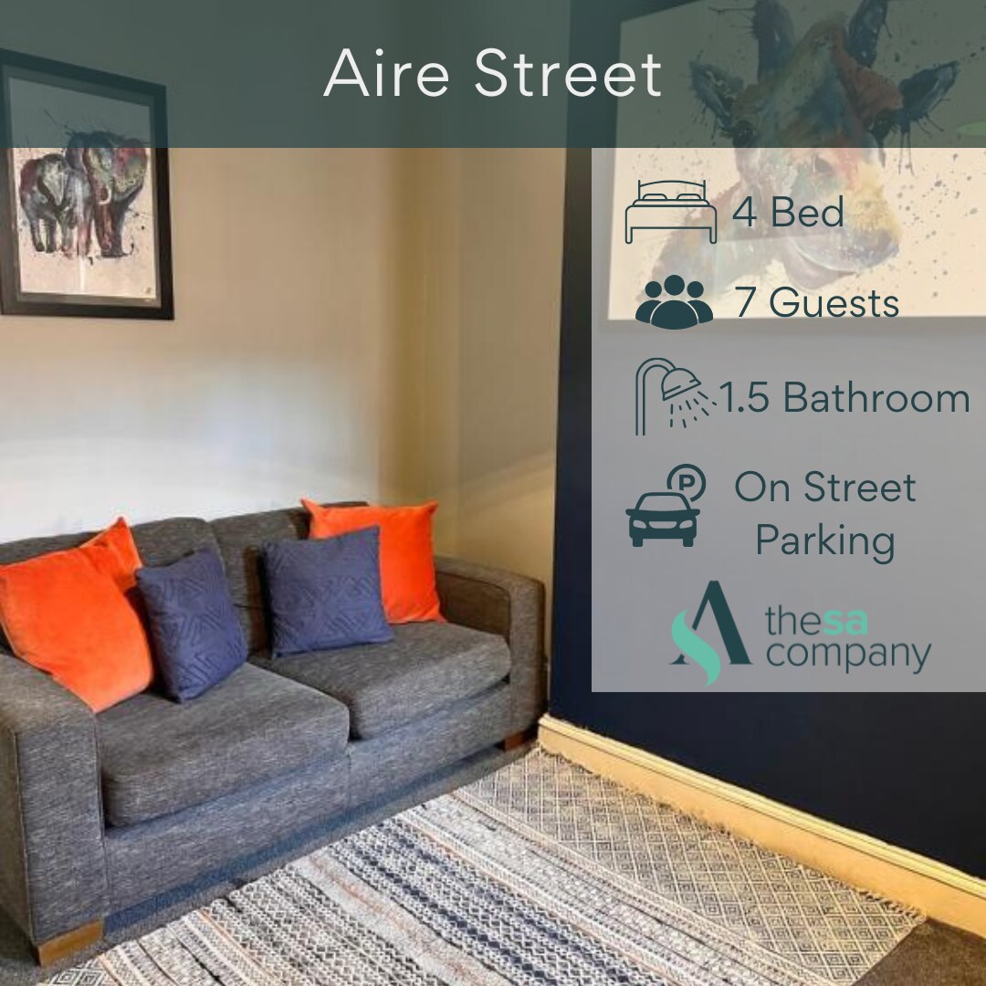 A comfortable seating area features a dark gray sofa adorned with orange and blue throw pillows. Colorful artwork is displayed on the walls, and a woven rug adds texture to the space. Natural light enhances the welcoming feel of the room.