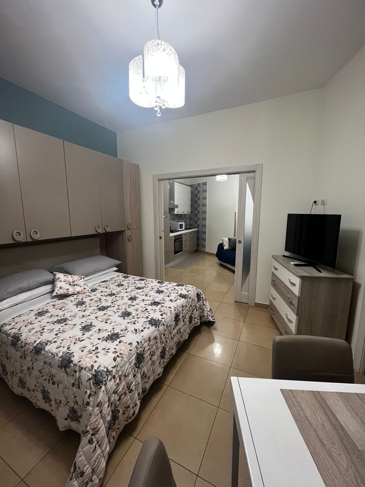 The sleeping area features a spacious bed adorned with a floral-patterned coverlet. A television is positioned on a light-colored dresser, while a small dining table is visible. An archway leads into the kitchen area, showcasing a modern setup with appliances in soft colors.