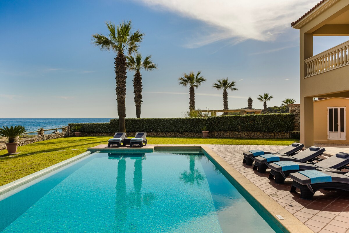 A serene outdoor area showcases a sparkling swimming pool surrounded by sun loungers. Palm trees line the background, providing a tropical feel. The coastline can be seen in the distance, with a clear blue sky overhead, enhancing the peaceful atmosphere.