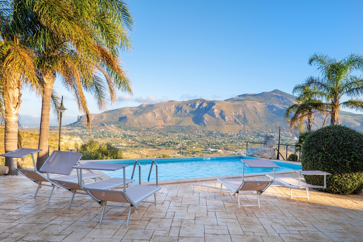An inviting pool area features several lounge chairs arranged around a tranquil swimming pool. Lush palm trees provide shade, while panoramic mountain views create a serene backdrop, enhancing the space's natural beauty.
