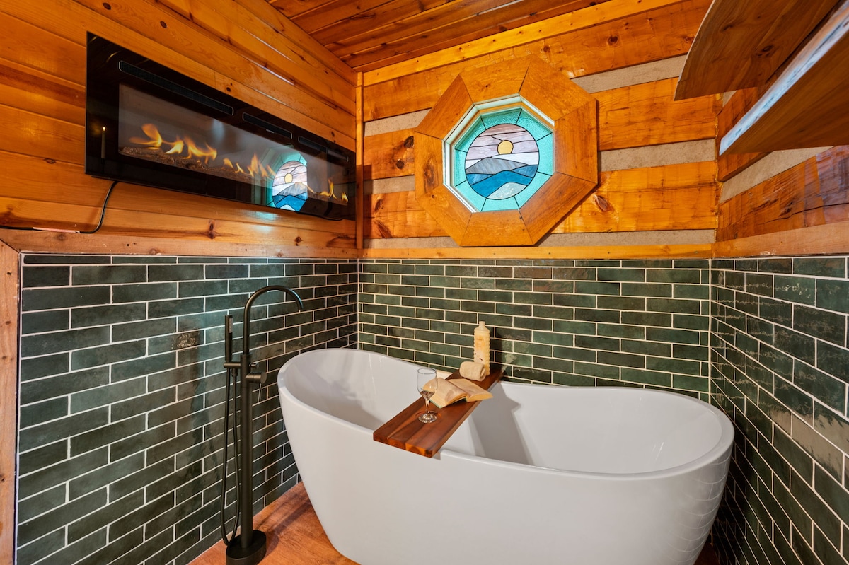 A luxurious soaking tub is centered in the room, complemented by a wooden bath tray. A stained glass window provides natural light, while a modern fireplace is mounted on the wall, adding a cozy element to the rustic wood-paneled space.