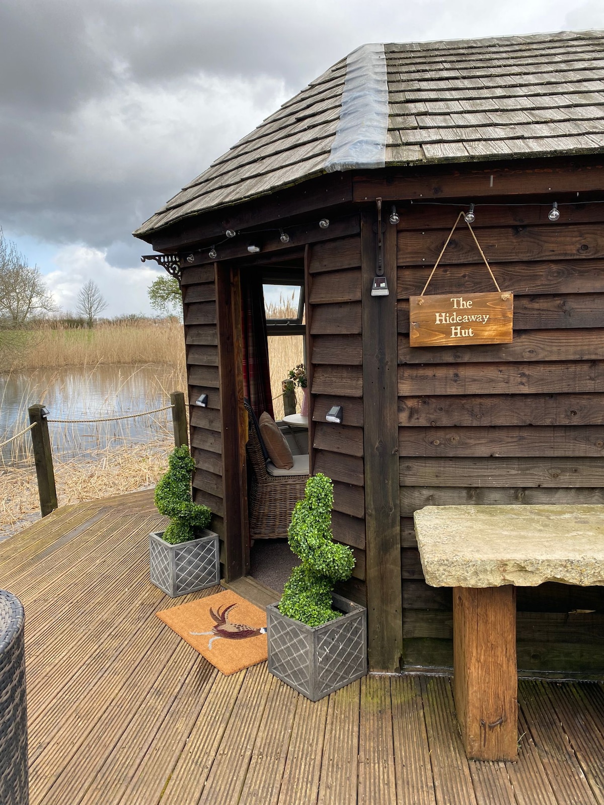The exterior of the hideaway hut is shown, featuring rustic wooden siding and a charming entrance with a welcome mat. Potted greenery flanks the doorway, and a stone table is present on the deck. A serene pond with reeds is visible in the background.