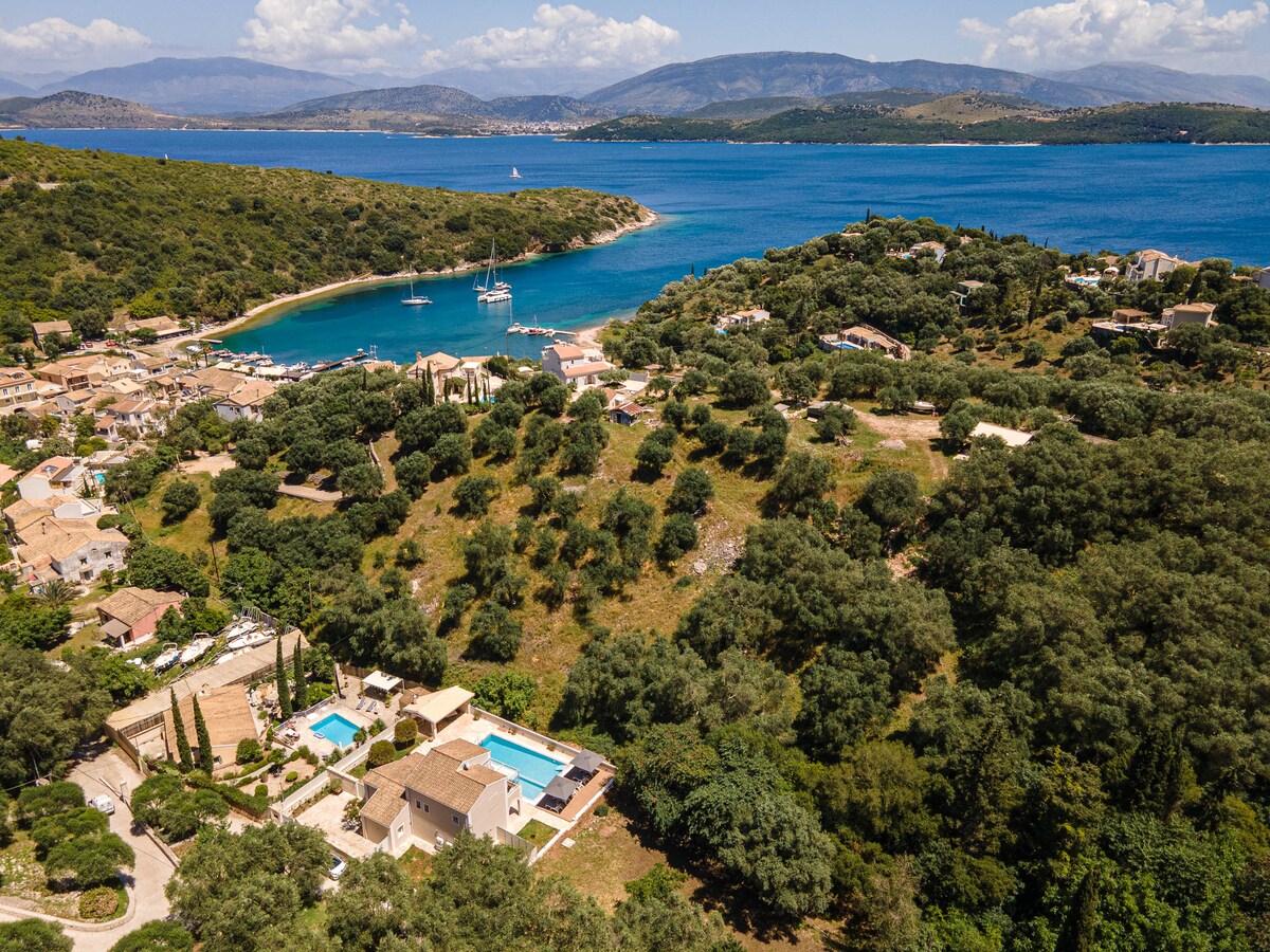 This aerial view captures the villa's expansive grounds amidst lush olive groves, with two private pools visible. The vibrant blue waters of Agios Stefanos Bay are framed by green hills, showcasing the peaceful coastal surroundings. Nearby boats can be seen moored along the shore.