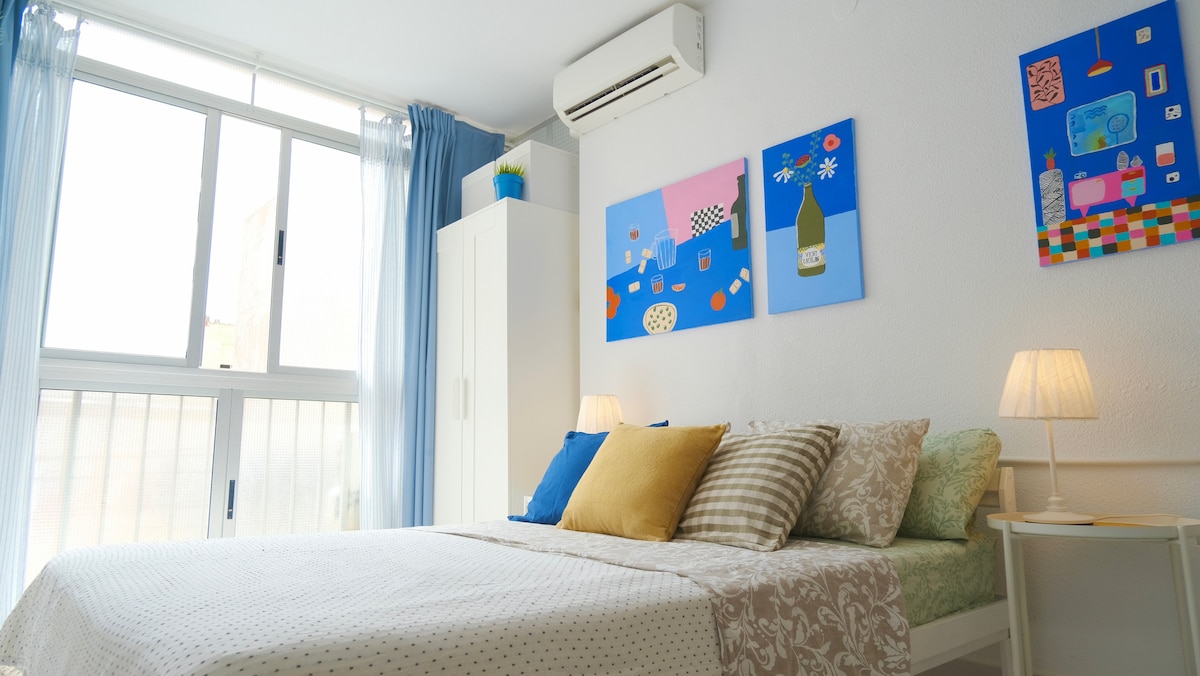 The inviting bedroom features a spacious bed adorned with a patterned cover and decorative pillows. Two pieces of colorful wall art brighten the room. Natural light floods through the large window, complemented by sheer curtains. A nightstand holds a lamp, enhancing the cozy ambiance.