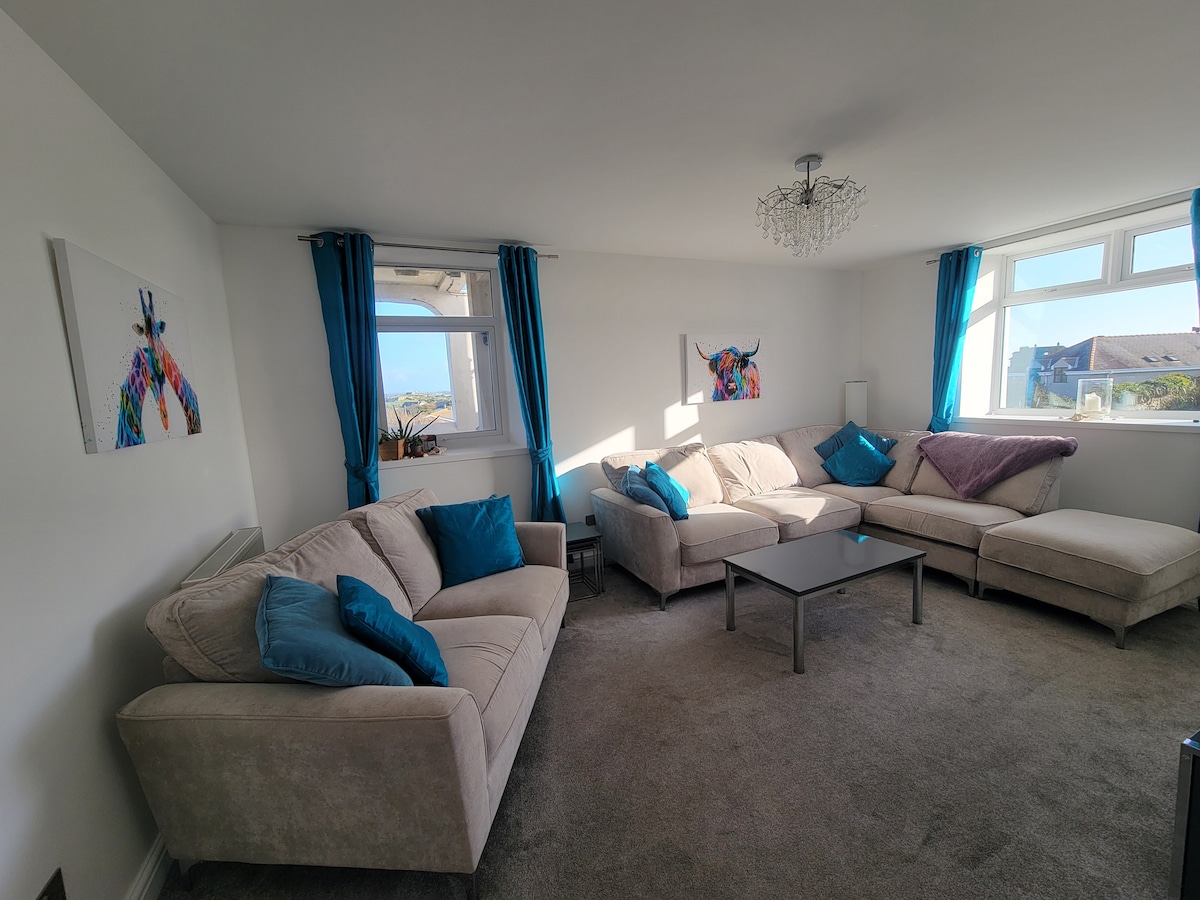 A light-filled lounge area features a large L-shaped sectional sofa with accent pillows in shades of blue. A separate two-seater sofa complements the space. Two windows allow natural light to filter in, while artwork highlights the walls. A central coffee table sits on soft carpet.