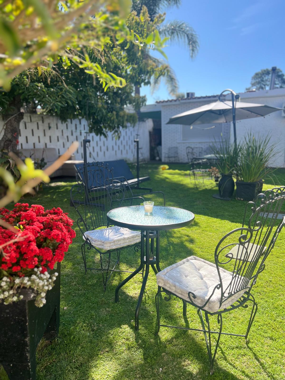 A charming outdoor space features a small round table surrounded by two wrought-iron chairs, set on a lush green lawn. Bright red flowers bloom in planters nearby, while an umbrella provides shade in the background, creating a tranquil atmosphere.