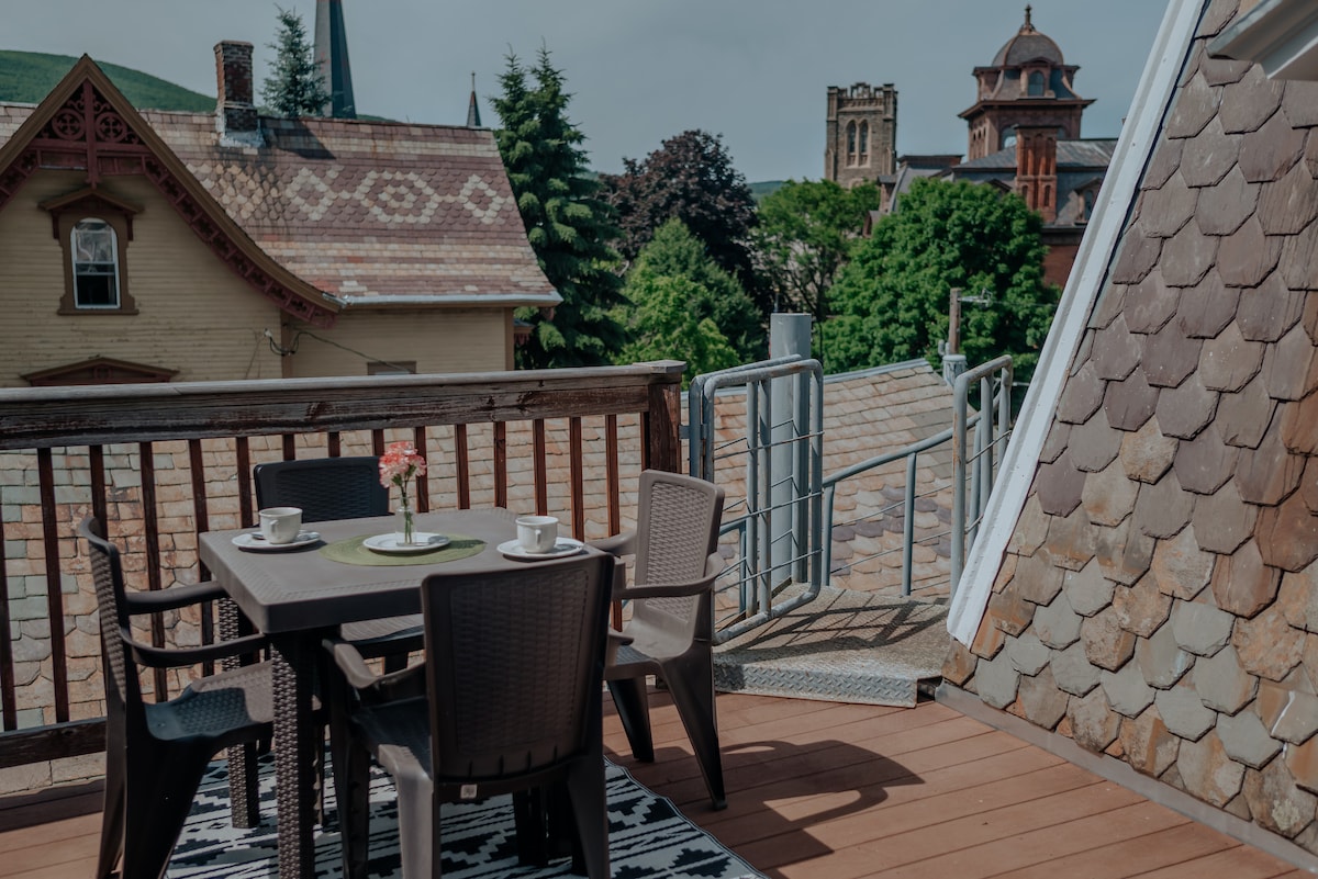 A balcony deck is displayed with a dining table, four chairs, and a vase containing flowers. The surrounding rooftops and greenery create a scenic view, with a historic building visible in the background under a clear sky.