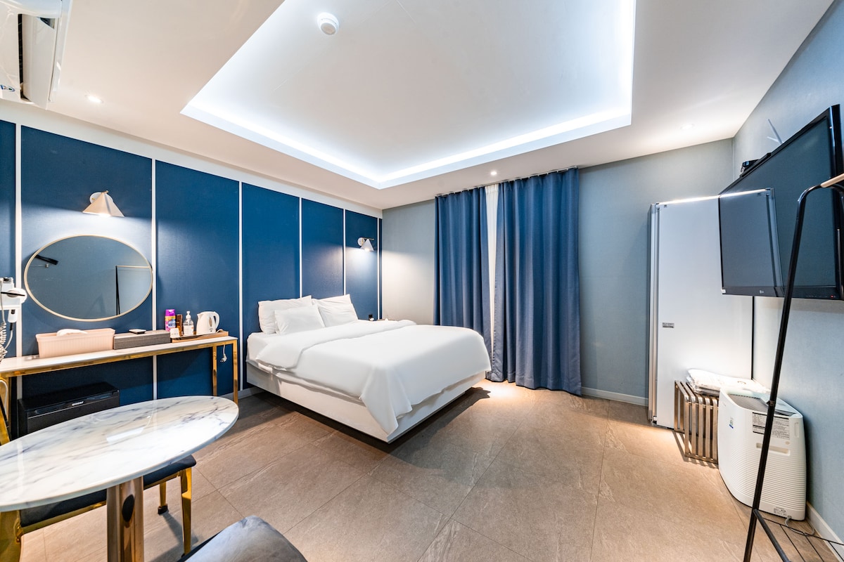 A spacious hotel room is presented with a large bed against a blue accent wall. A round mirror and desk are positioned nearby, complemented by a dining table. Floor-to-ceiling curtains frame a window, while a refrigerator and television are placed along the opposite wall.