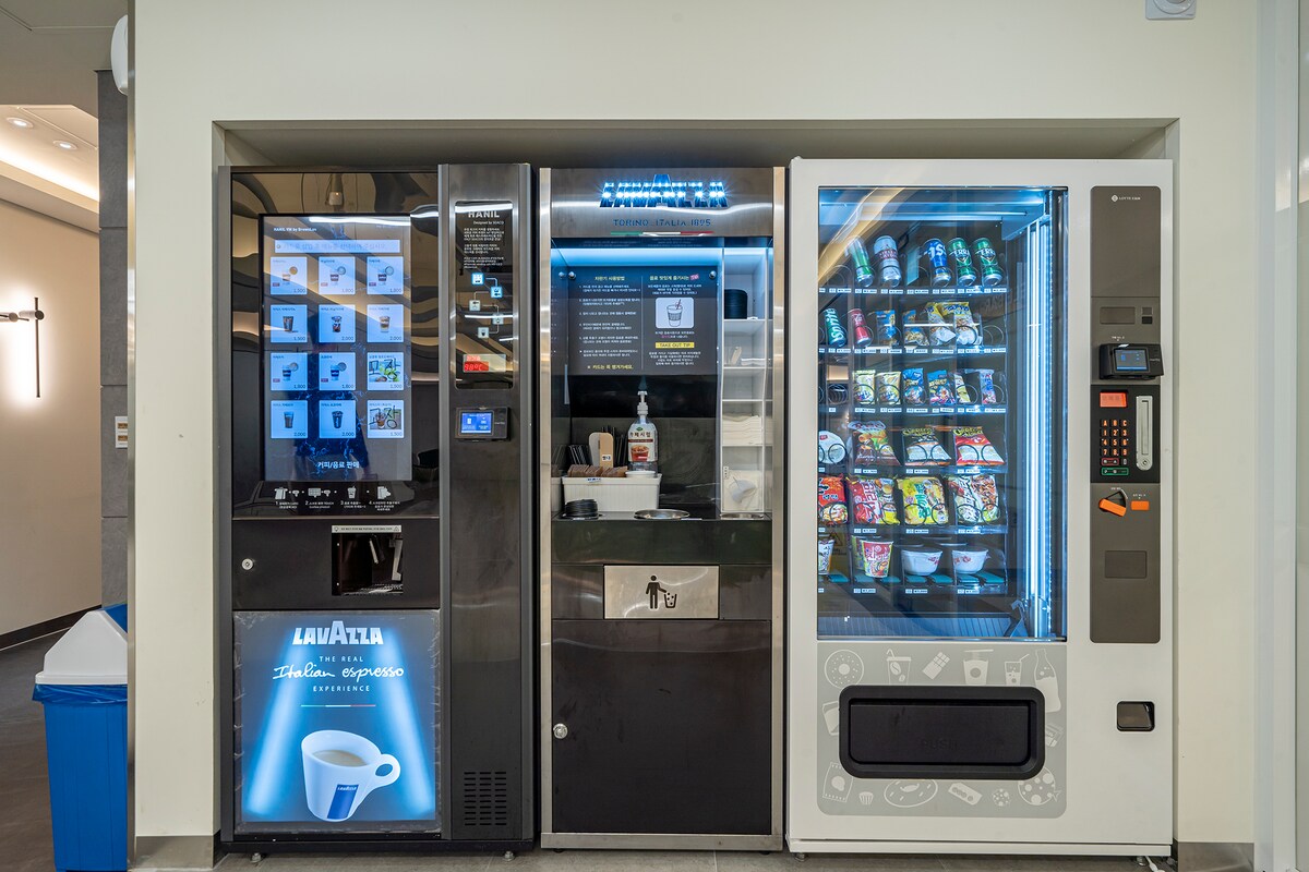 A vending area features three machines offering a variety of snacks and beverages. The left machine displays coffee options, including an espresso maker, while the center machine contains bottled drinks. The right machine is filled with an assortment of packaged snacks, all illuminated for visibility.