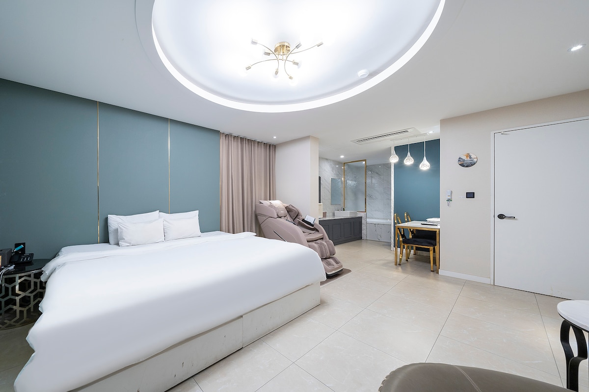 A spacious guest room features a large bed adorned in crisp white linens. Decorative light fixtures hang from the ceiling, while soft blue walls create a calming atmosphere. A massage chair is positioned nearby, and a stylish desk with chairs is visible in the background.