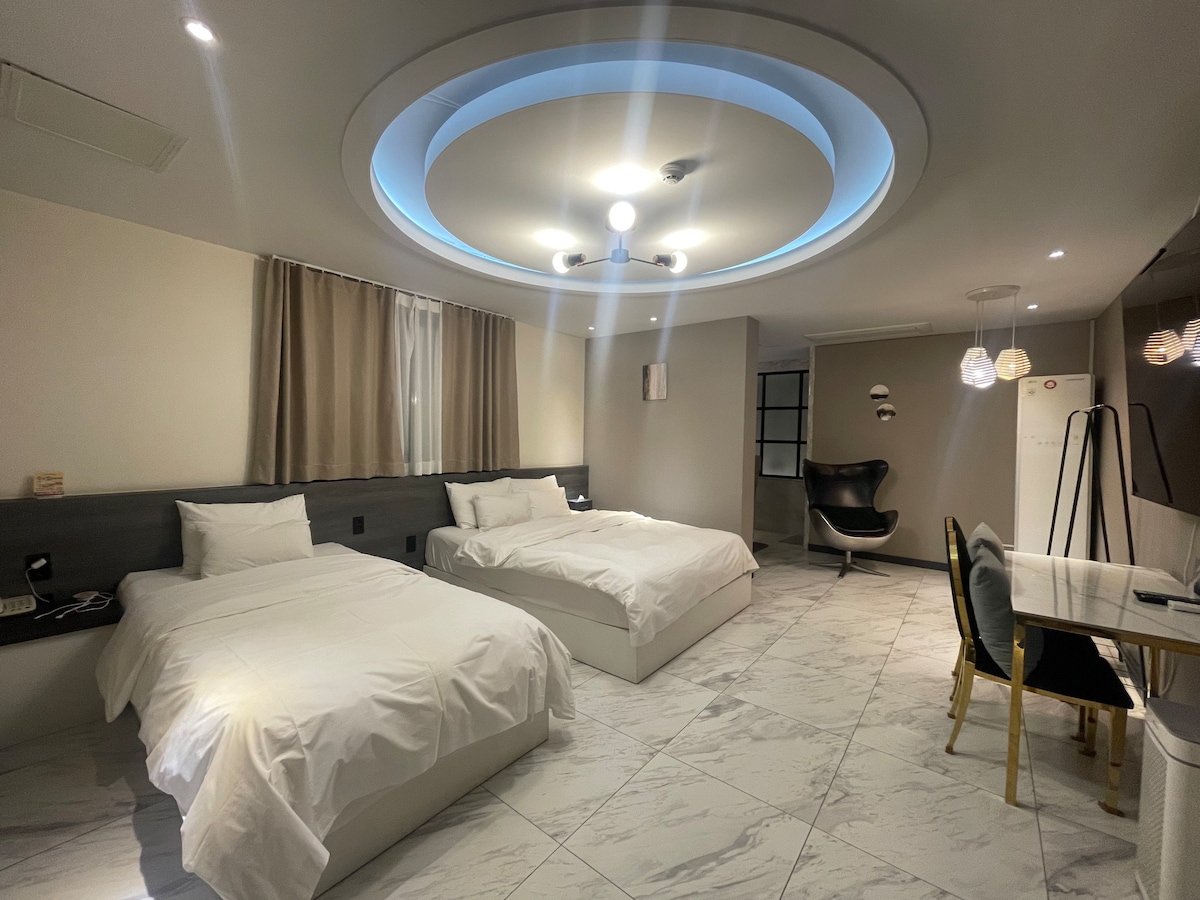 A spacious room contains two large beds with crisp white bedding, complemented by soft, neutral-colored curtains. Modern lighting features a circular design on the ceiling, illuminating the area. A dining table and chairs are positioned near a stylish armchair, creating an inviting seating arrangement.