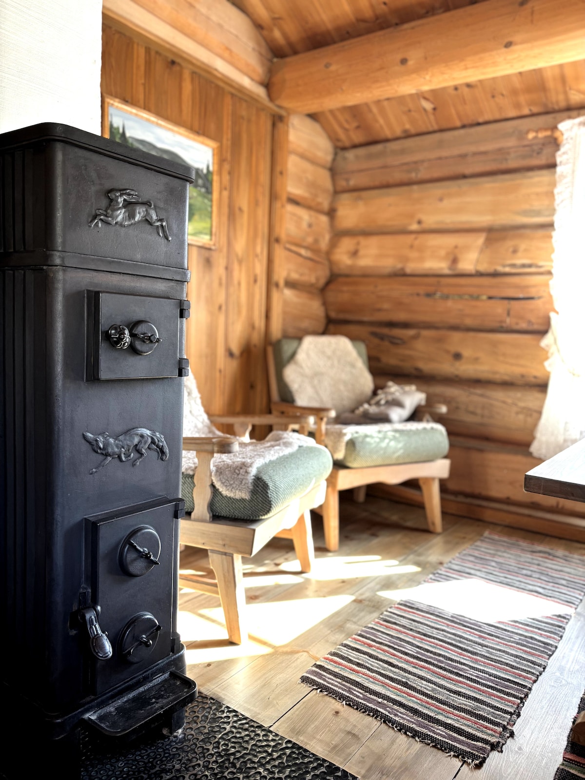 A charming corner of the interior features a traditional wood-burning stove, crafted with intricate designs. Two comfortable armchairs with soft throws are positioned nearby. A striped rug covers the floor, enhancing the cozy rustic atmosphere of the log cabin setting.