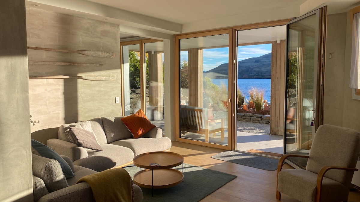 A light-filled living area features large glass doors that open to a scenic view of the lake, allowing for easy access to the outdoor space. The room includes a cozy seating arrangement with soft grey sofas, a circular wooden coffee table, and a gentle color palette.
