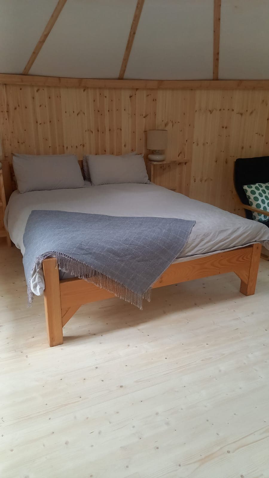 A spacious bed is situated in a wooden yurt, featuring soft gray and beige bedding. Two plump pillows rest at the head of the bed, while a knitted throw is casually draped across the foot. Natural wood walls enhance the cozy ambiance.