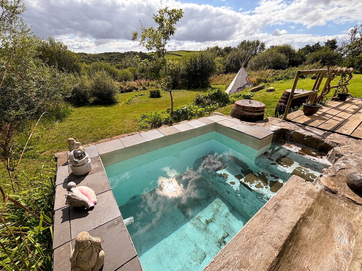 A natural plunge pool is set within stone borders, filled with clear water reflecting the sky. Surrounding greenery and a view of a teepee create a serene outdoor setting. A wooden deck with seating areas complements the landscape, inviting relaxation.