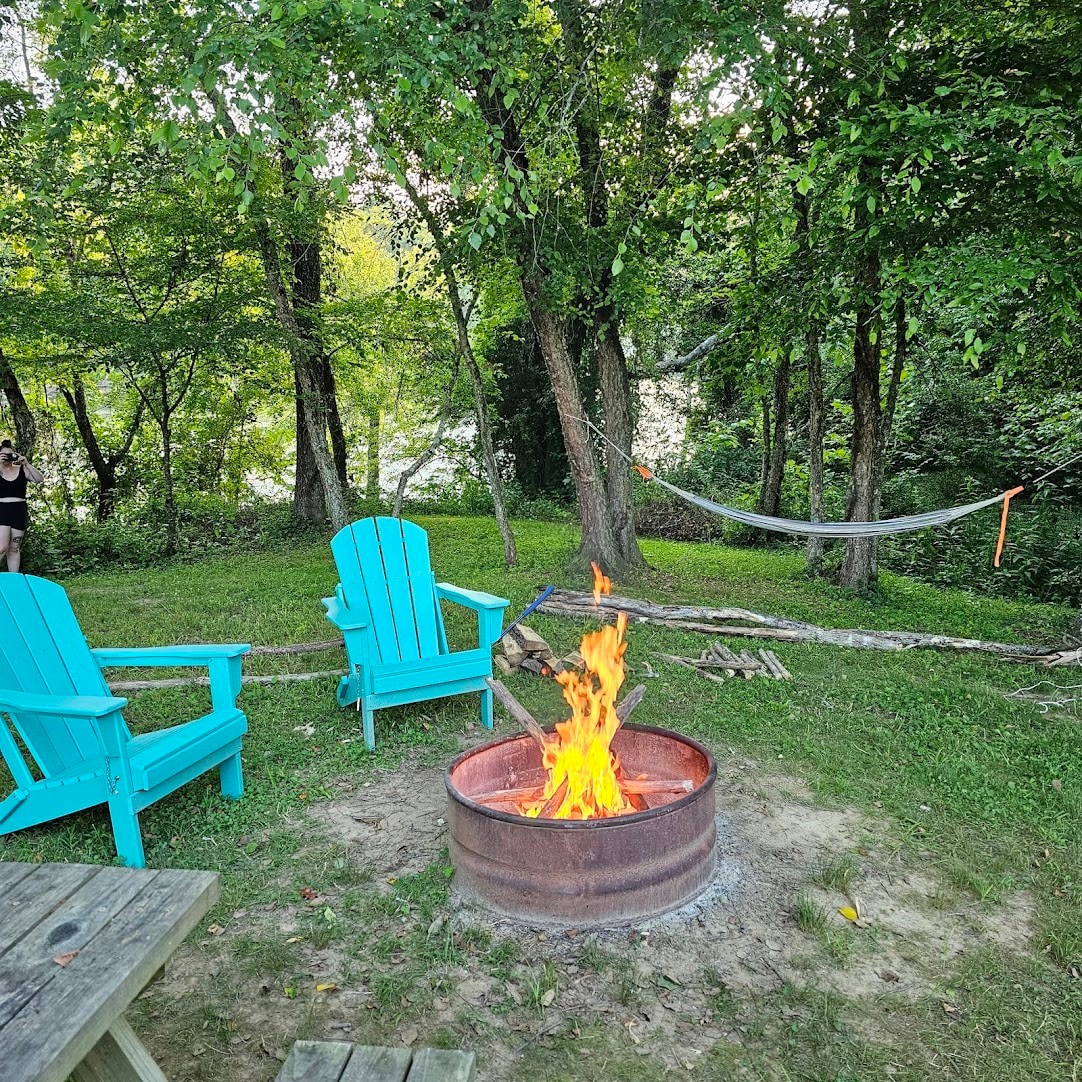 A fire ring is centrally located, with a lively fire burning bright. Two turquoise adirondack chairs are positioned nearby, surrounded by lush greenery. A hammock is visible in the background, offering a serene spot for relaxation.