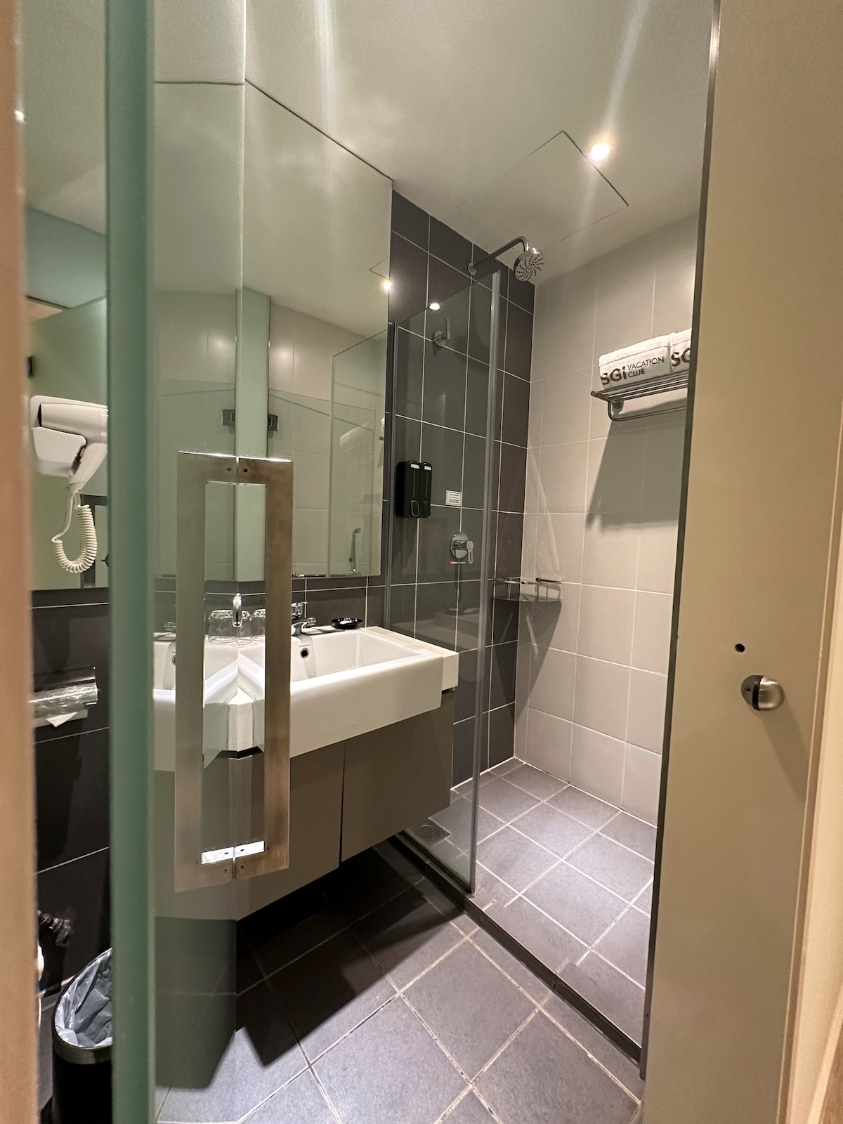 A modern bathroom is highlighted by a spacious walk-in shower enclosed by glass panels. The sink area features a contemporary vanity with a mirror above. Grey tiled flooring complements the neutral tones of the walls, enhancing the sleek design.