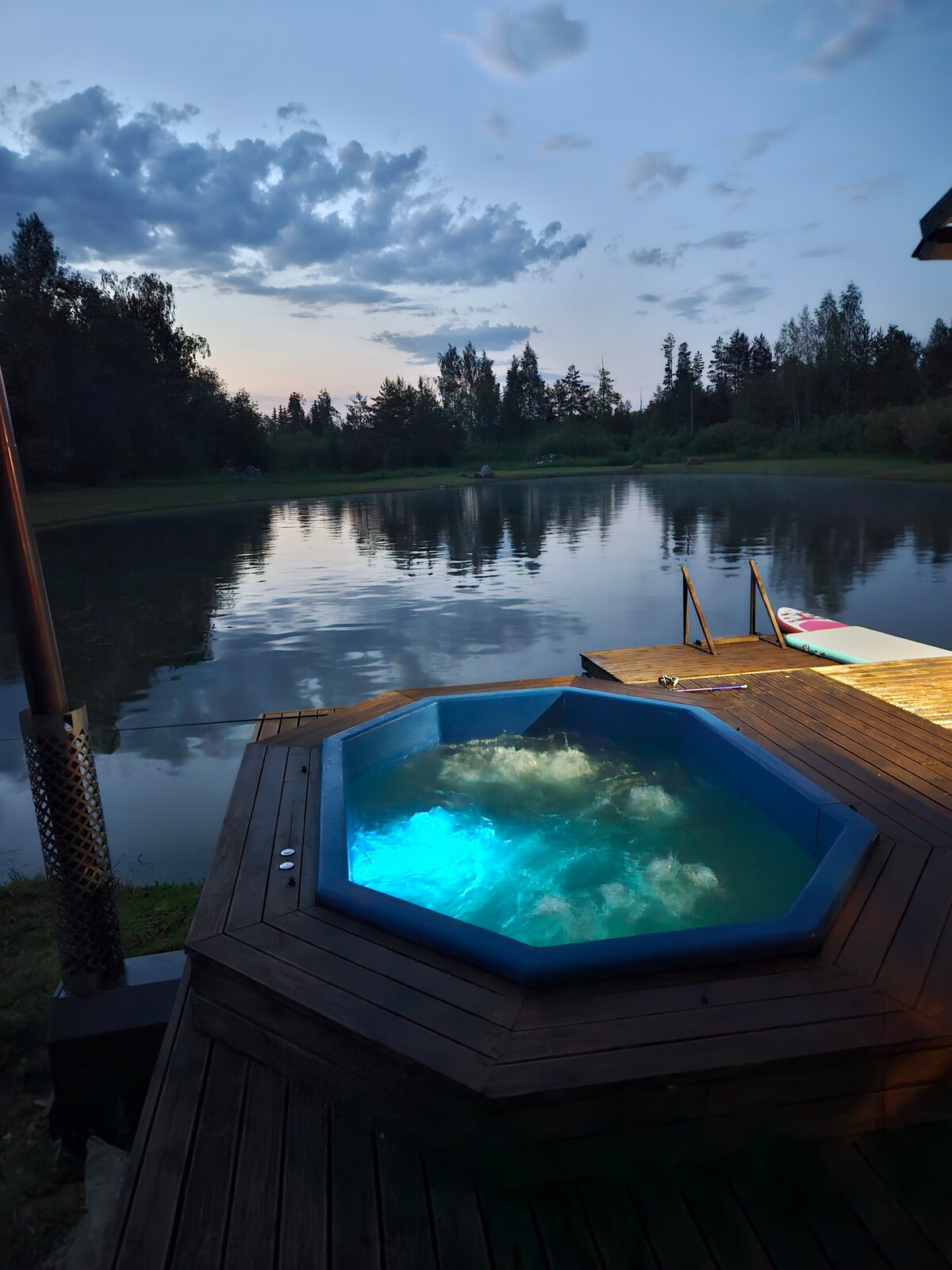 A private hot tub sits on a wooden deck, surrounded by lush greenery and reflecting in a calm body of water. The evening sky presents soft clouds, while serene blue lighting illuminates the water's surface.