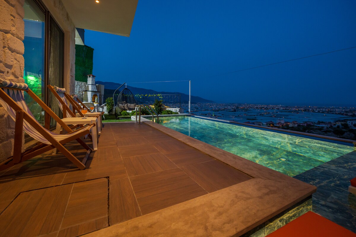 An outdoor terrace features a serene infinity pool with views of the surrounding landscape under a night sky. Lounge chairs are positioned nearby, inviting relaxation. Soft lighting creates a calming atmosphere, enhancing the tranquil setting.