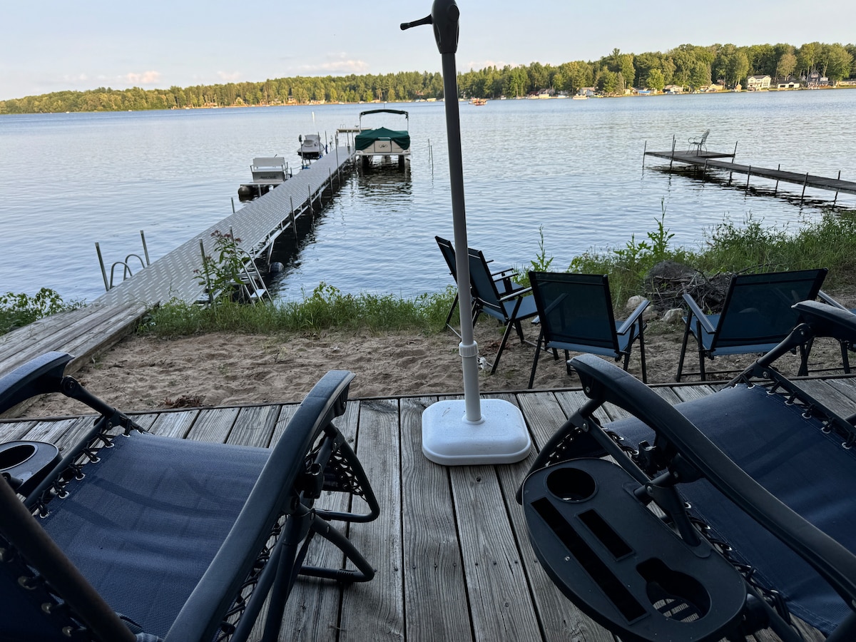 A serene lakeside view is displayed, featuring a wooden patio area with two lounge chairs. A dock extends into the calm waters of Rose Lake, with several boats visible in the distance. Lush greenery frames the shoreline, enhancing the natural beauty of the setting.